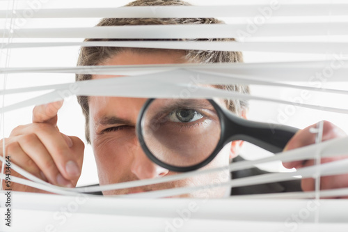 Businessman peeking through blinds with magnifying glass