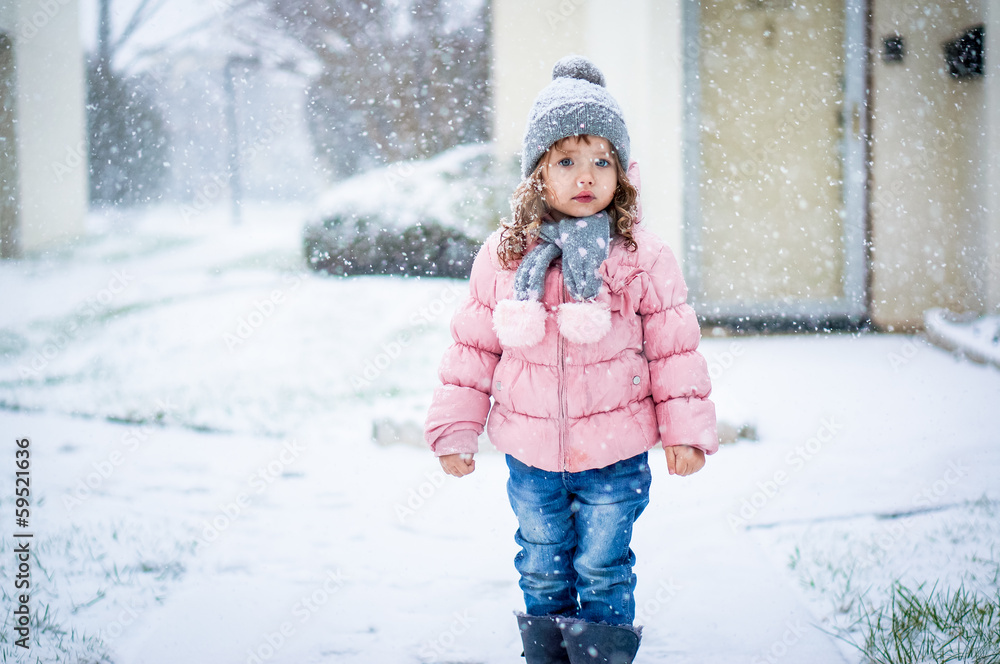 Cute baby girl in pink jacket and grey hat enjoying first snow b
