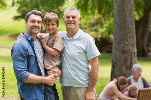 Grandfather, father and son with family in background at park