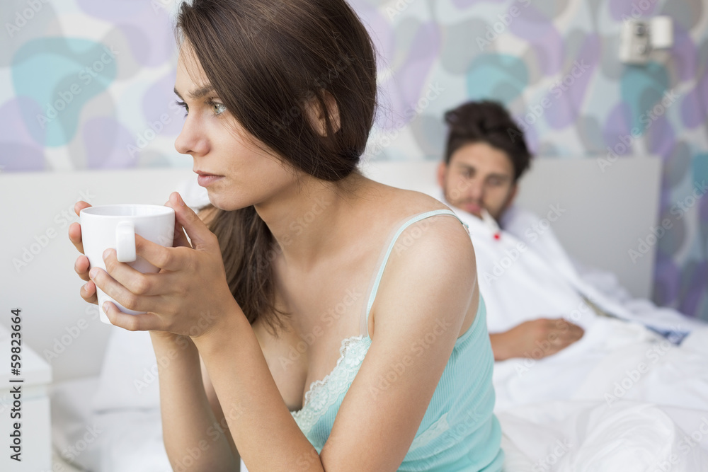 Worried young woman holding coffee mug while ill man relaxing in background Worried young woman holding coffee mug while ill man relaxing in background
