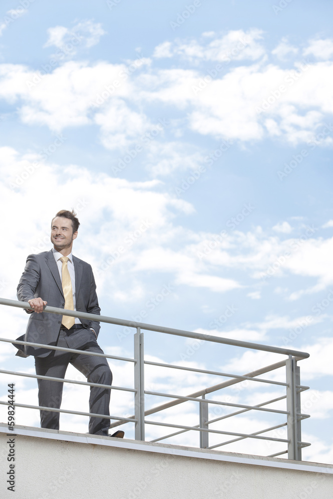 Young businessman standing at terrace railings against sky Young businessman standing at terrace railings against sky