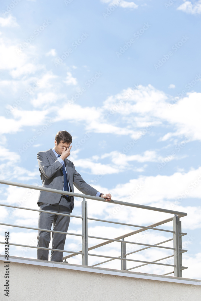 Stressed young businessman rubbing eyes on terrace against sky