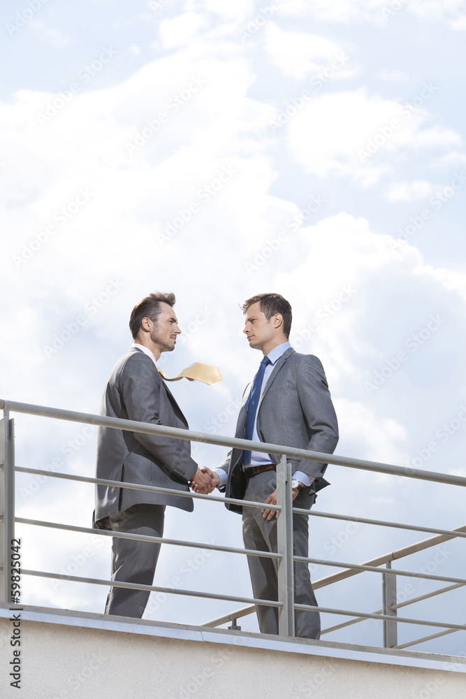 Side view of businessmen shaking hands at terrace railings against sky Side view of businessmen shaking hands at terrace railings against sky