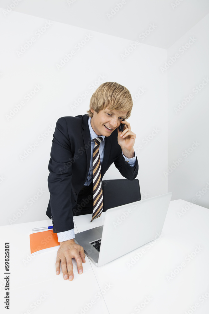 Happy businessman using cell phone while looking at laptop in office