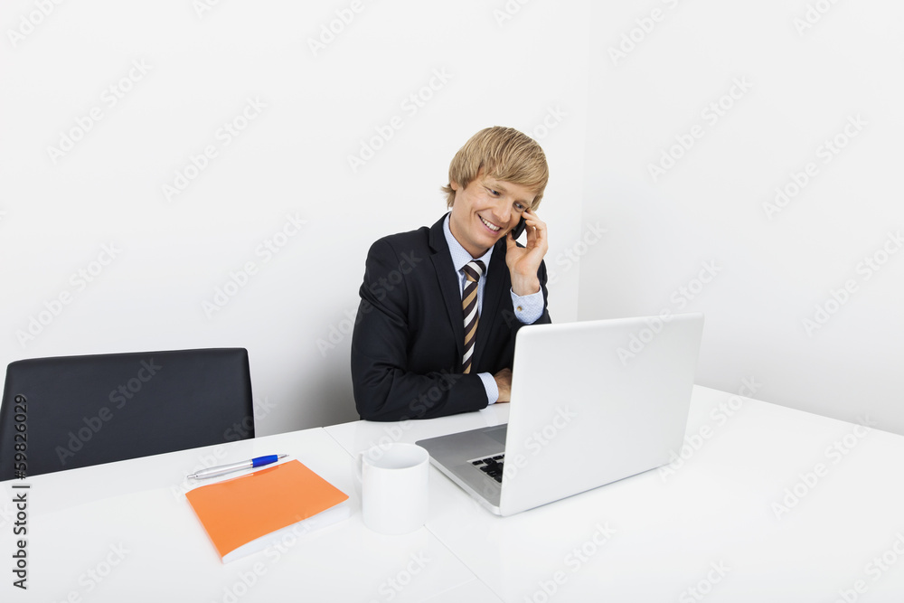 Happy mid adult businessman on call using laptop at desk in office Happy mid adult businessman on call using laptop at desk in office
