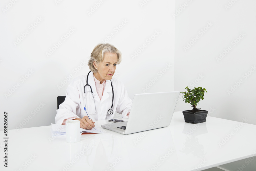 Senior female doctor writing notes while looking at laptop in clinic Senior female doctor writing notes while looking at laptop in clinic