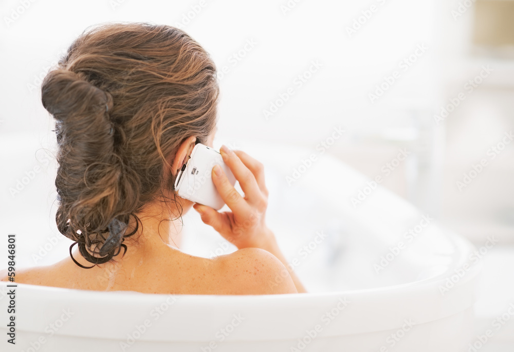 Young woman in bathtub talking cell phone. rear view - obrazy, fototapety, plakaty 