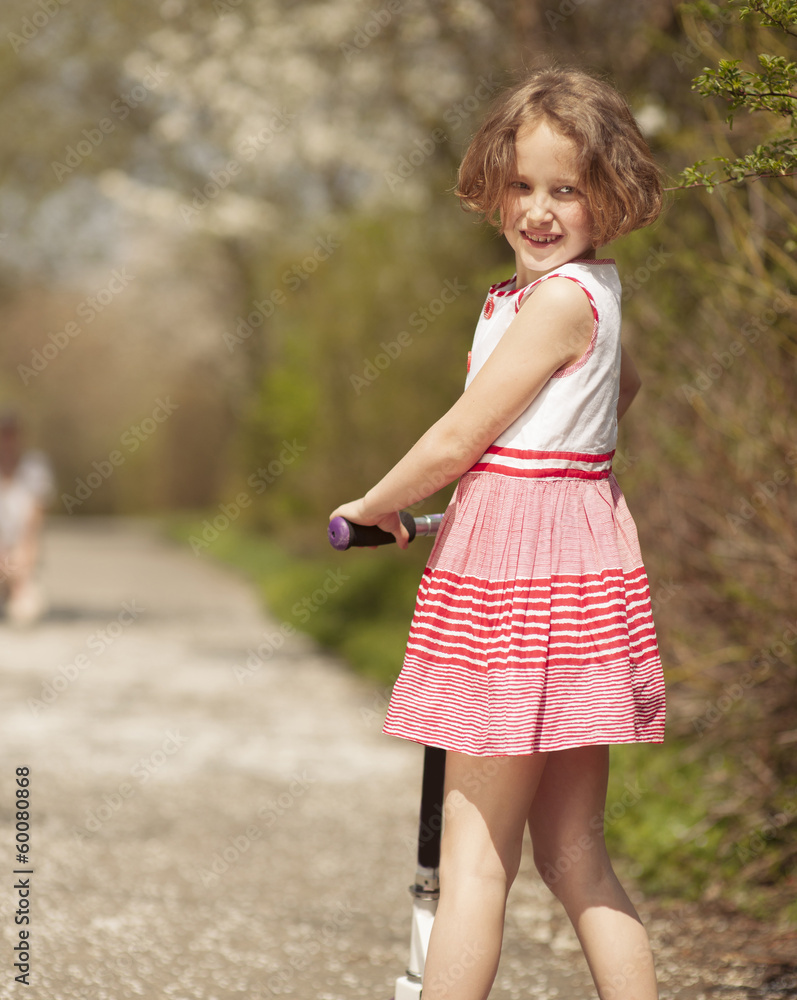 Young girl riding scooter in park away from camera to mother Young girl riding scooter in park away from camera to mother
