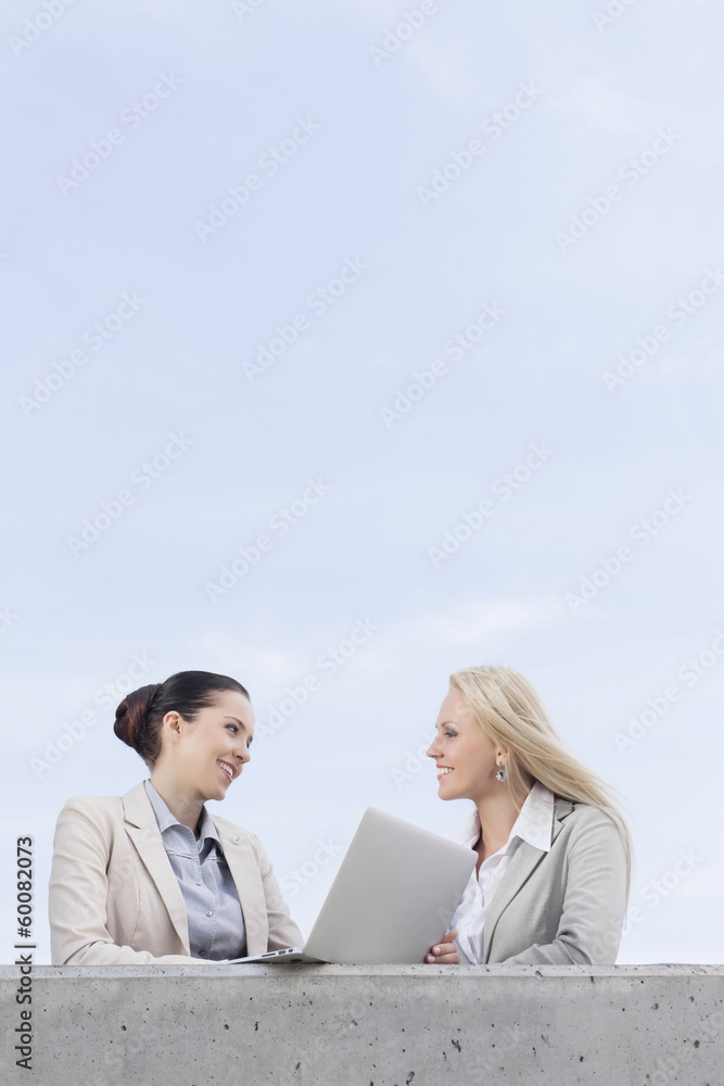 Low angle view of happy young businesswomen with laptop discussing while standing on terrace against sky