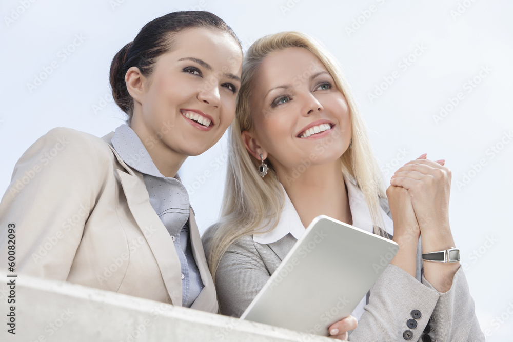 Low angle view of happy businesswomen with digital tablet looking away against clear sky Low angle view of happy businesswomen with digital tablet looking away against clear sky