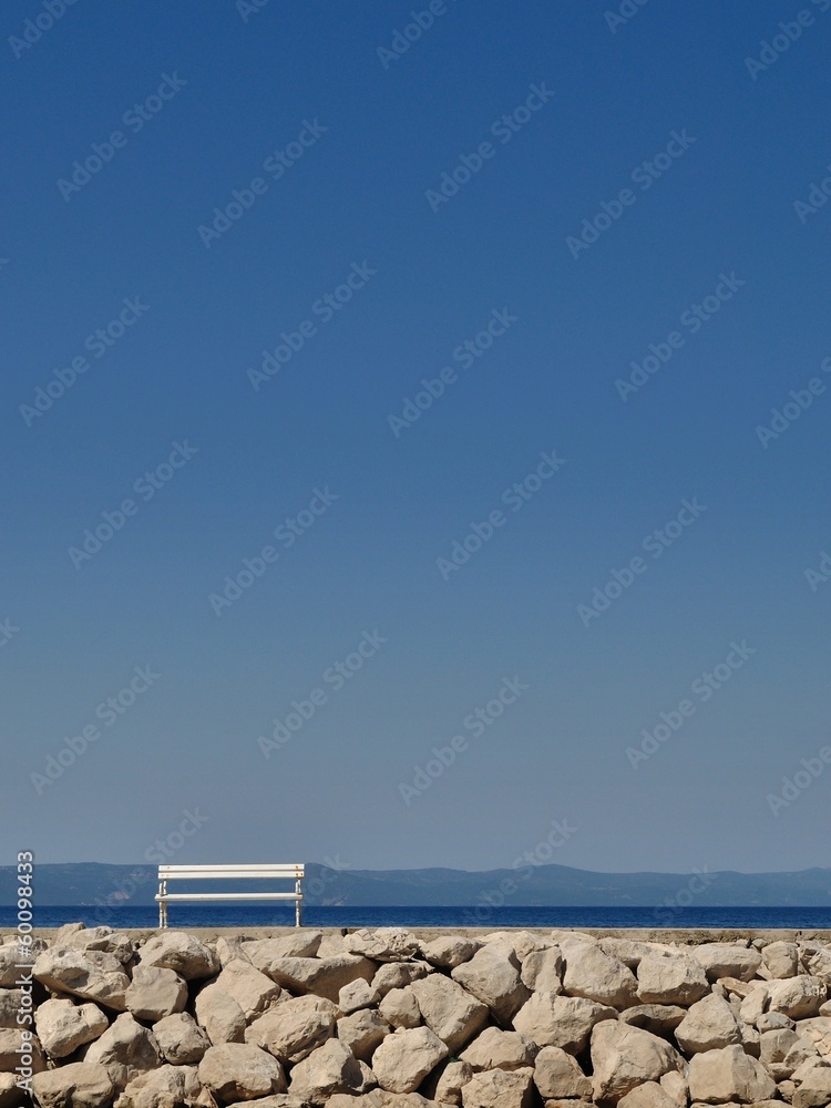 White bench in port with stones and blue sky. Podgora, Croatia