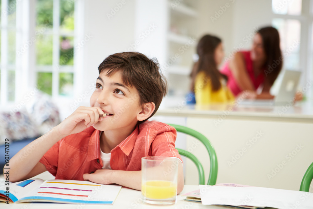 Boy Doing Homework As Mother And Daughter uses Laptop