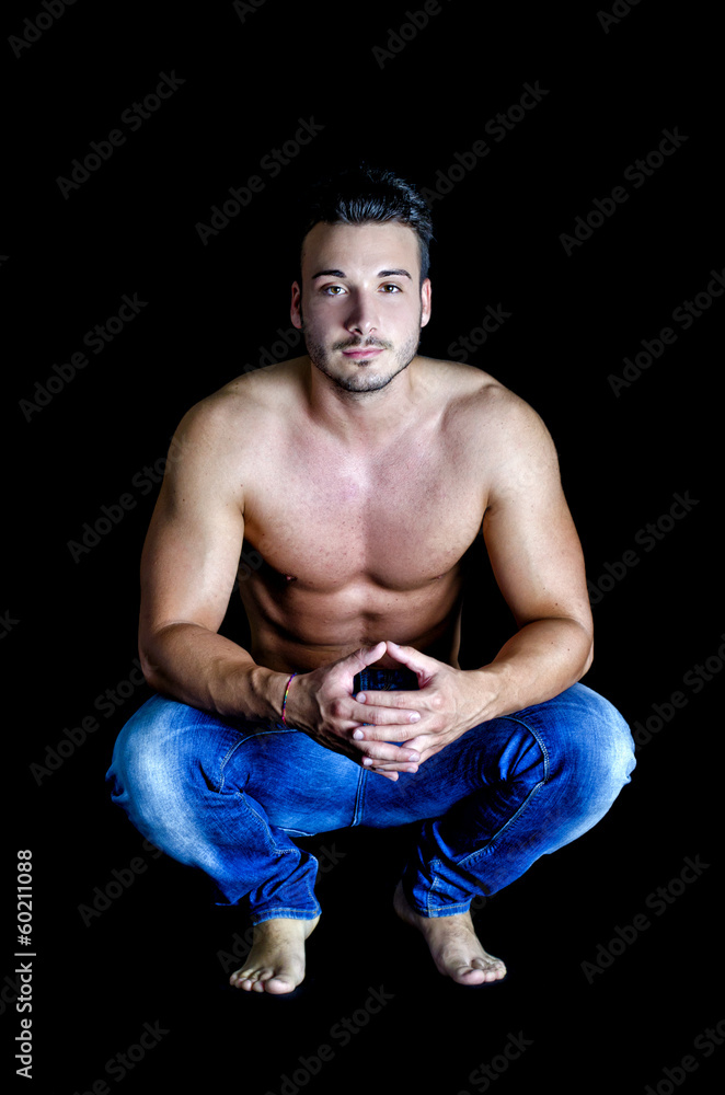 Shirtless muscular young man in jeans sitting on dark background Shirtless muscular young man in jeans sitting on dark background