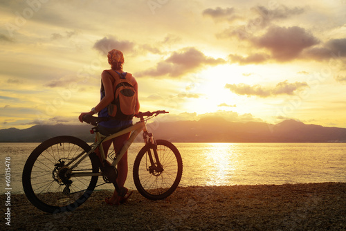 Young woman with backpack standing on the shore near her bicycle