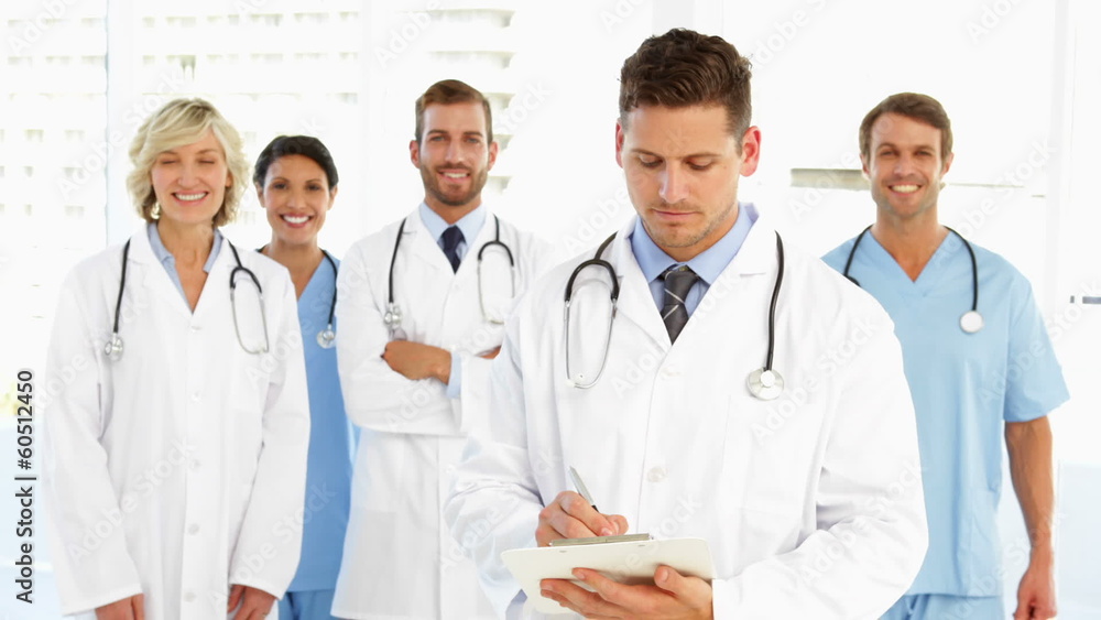 Serious doctor writing on clipboard while staff are behind him Serious doctor writing on clipboard while staff are behind him