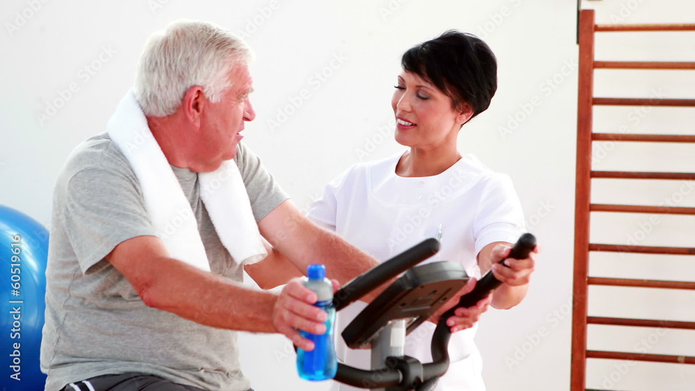Elderly man using the exercise bike talking to physiotherapist Elderly man using the exercise bike talking to physiotherapist