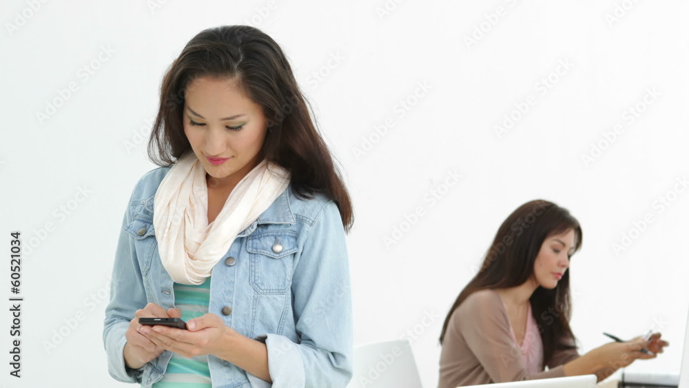 Creative businesswoman sending a text with coworker behind her Creative businesswoman sending a text with coworker behind her
