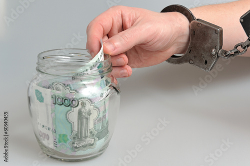 Рука в наручниках берет деньги из стеклянной банки