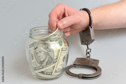 Рука в наручниках берет деньги из стеклянной банки