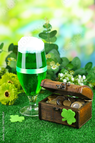 Glass of green beer and pitcher with coins on grass close-up