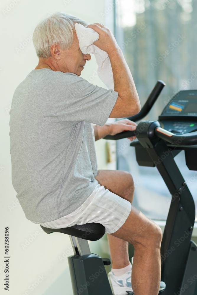 Tired senior man with towel on exercise bike in fitness club Tired senior man with towel on exercise bike in fitness club