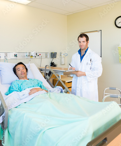 Doctor Writing On Clipboard By Patient's Bed In Hospital