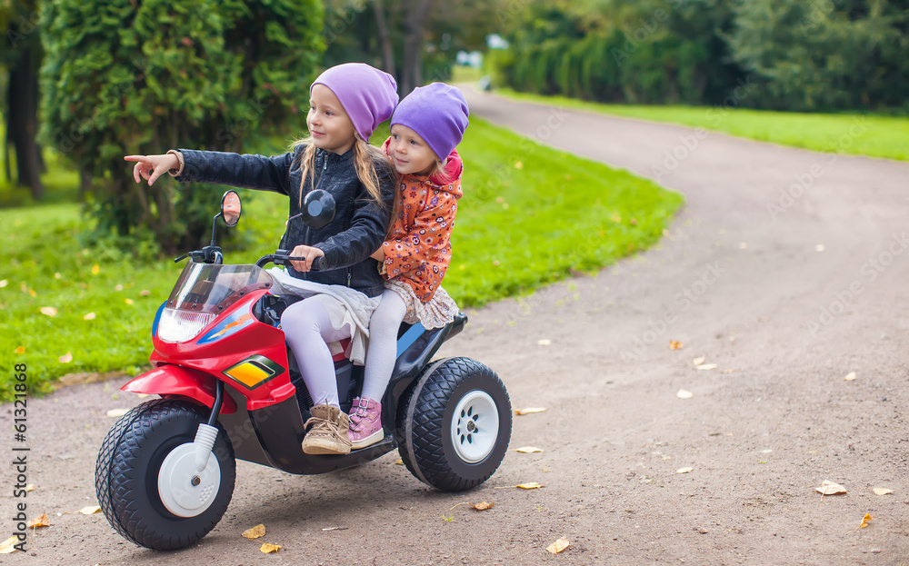 Little adorable sisters sitting on toy motorcycle in green park Little adorable sisters sitting on toy motorcycle in green park