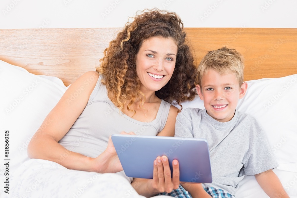 Smiling mother and son sitting on bed with tablet pc