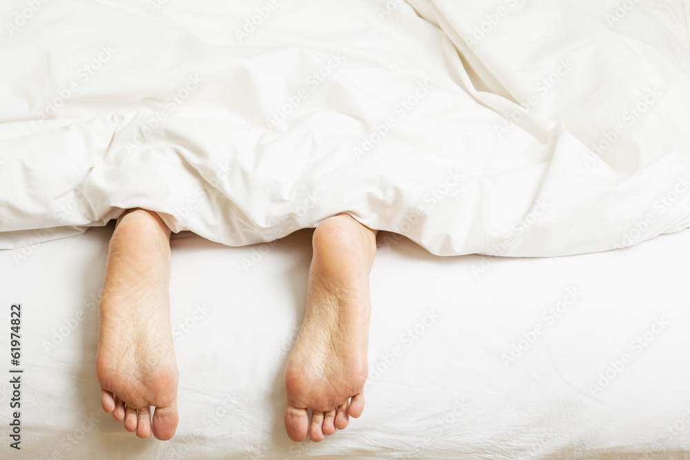 Woman's Feet Sticking Out Of Blanket On Bed At Home Woman's Feet Sticking Out Of Blanket On Bed At Home