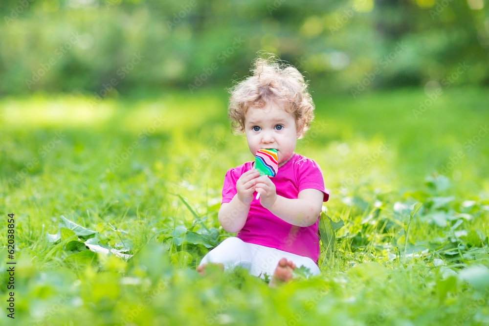 Adorable curly baby girl eating candy in a sunny summer park Adorable curly baby girl eating candy in a sunny summer park