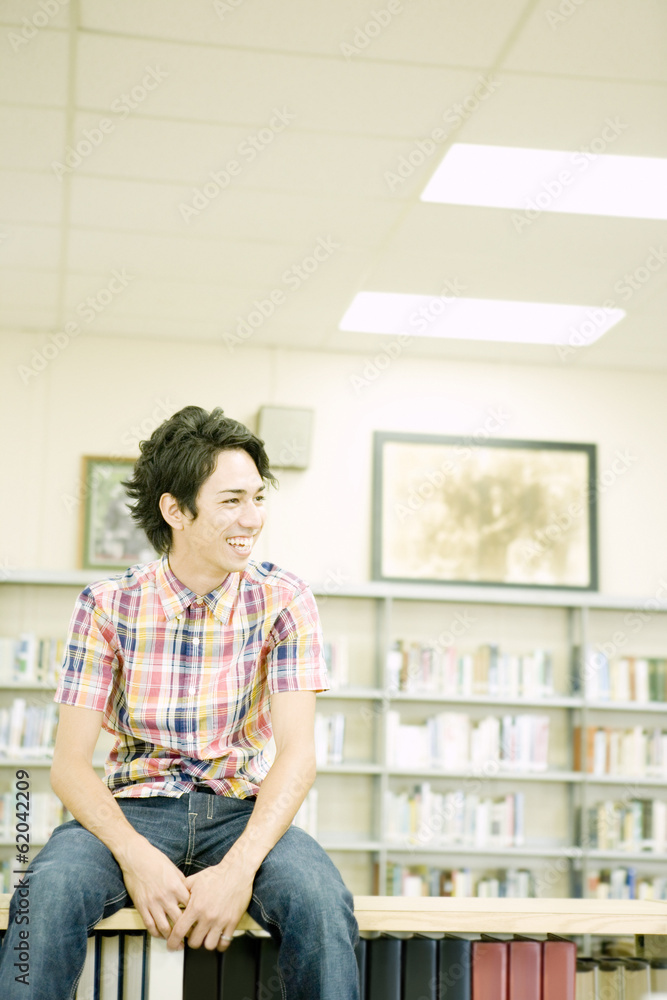 Fototapeta premium smiling male student sitting on bookshelf