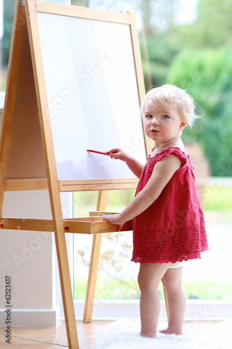 Toddler girl drawing on white board standing next window