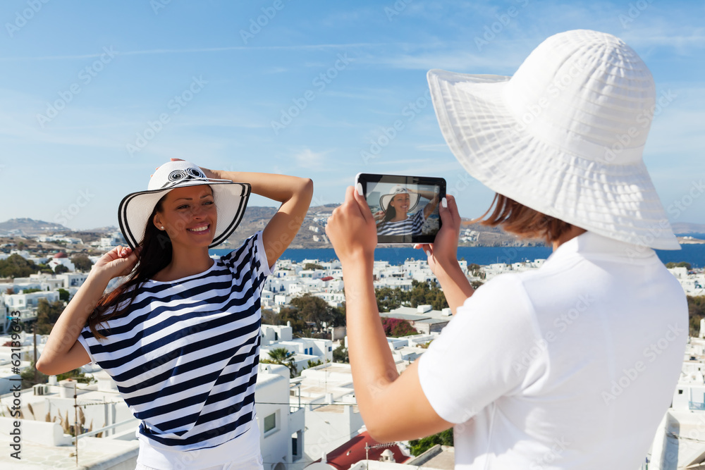 Fototapeta premium Young woman photographing her friend with a digital tablet. In a background is a beautiful Mykonos architecture.