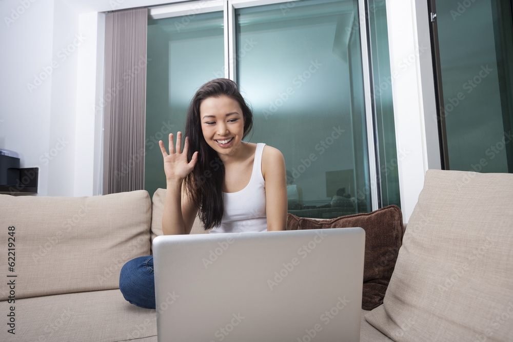 Naklejka premium Smiling young woman waving in front of laptop on sofa