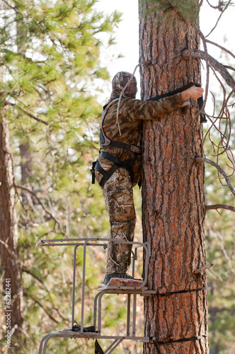Bow hunter in a ladder style tree stand attaching his safety harness above his head on the tree