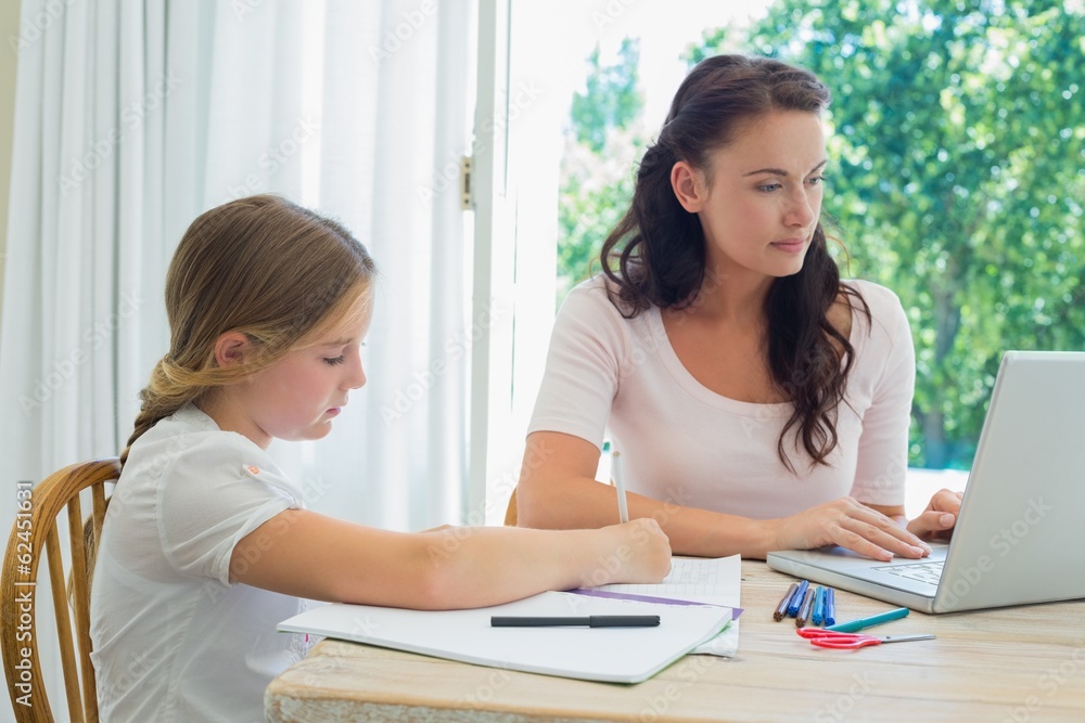 Girl doing homework while mother using laptop at table Girl doing homework while mother using laptop at table