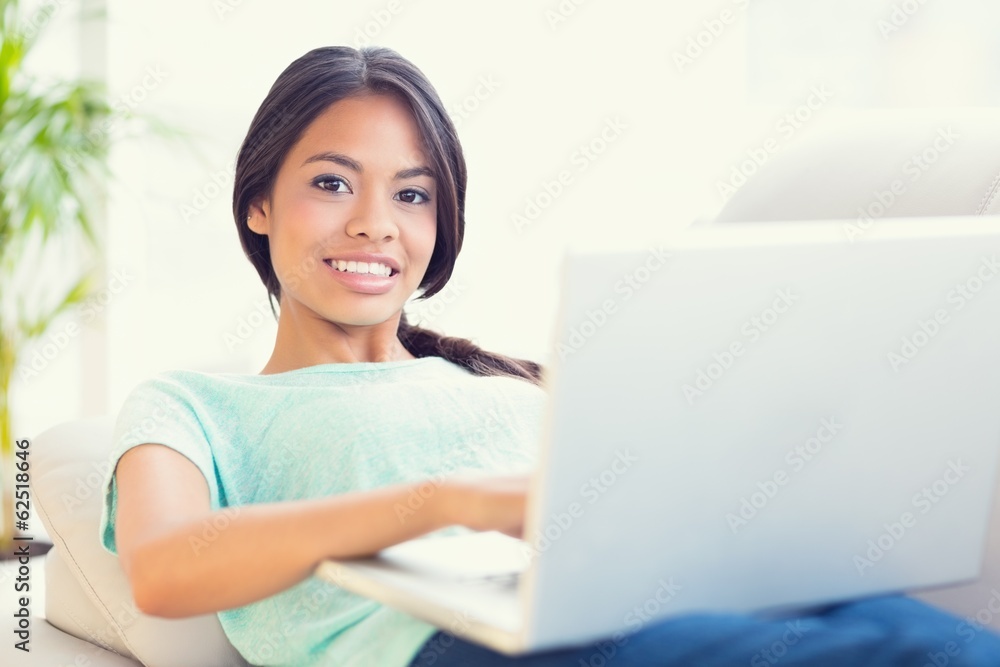 Happy girl lying on sofa using her laptop smiling at camera Happy girl lying on sofa using her laptop smiling at camera