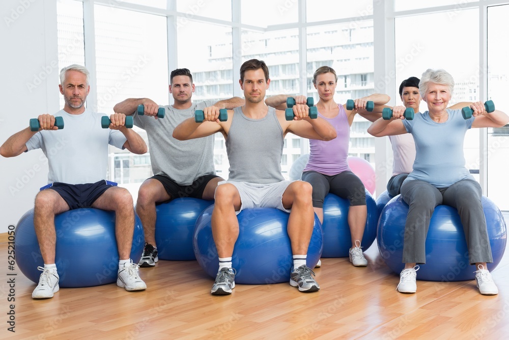 Fitness class with dumbbells sitting on exercise balls in gym Fitness class with dumbbells sitting on exercise balls in gym