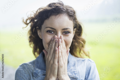 A young woman in a rural landscape, with windblown curly hair. Covering her face with her hands, and laughing.