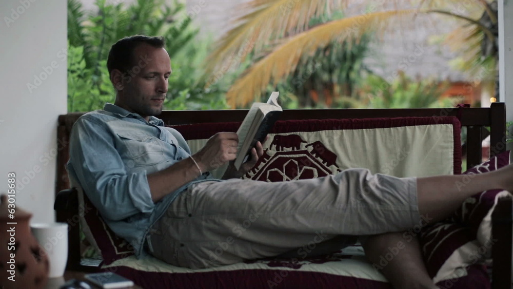 Young man on sofa reading book on patio Young man on sofa reading book on patio