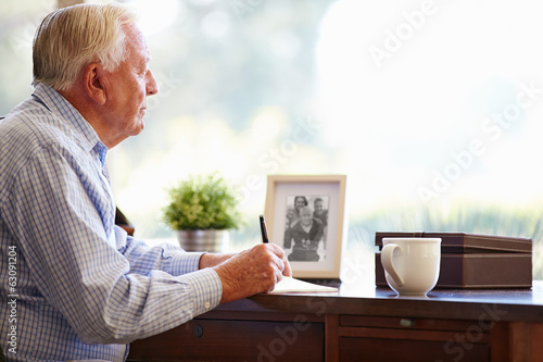 Senior Man Writing Memoirs In Book Sitting At Desk photo