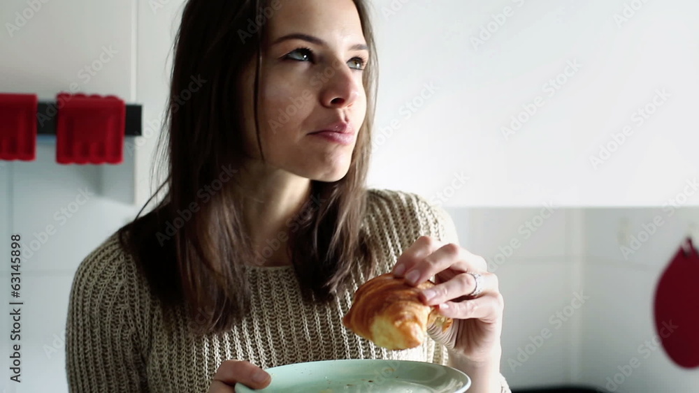 Young woman eating croissant in kitchen at home