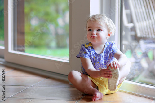 Little girl puts on her shoe sitting on the floor next window