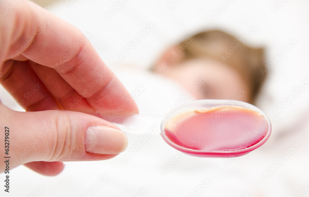 Mother holding medication in spoon foreground and sick little gi Mother holding medication in spoon foreground and sick little gi