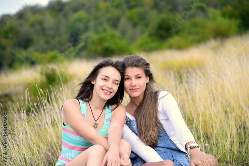 Two Beautiful Young Women Sitting On Dry Grass And Smiling