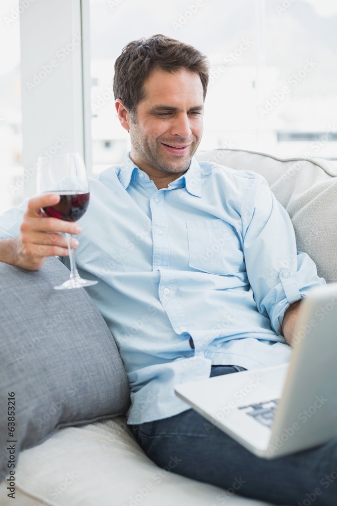 Happy man relaxing on sofa with glass of red wine using laptop Happy man relaxing on sofa with glass of red wine using laptop
