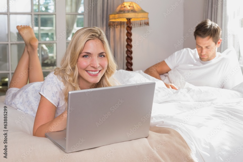 Woman using laptop while man reading book in bed Woman using laptop while man reading book in bed