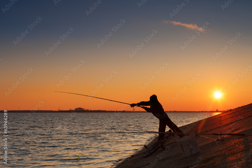 Silhouette of fisherman fishes on river bank against a sunset