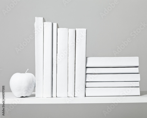 White Books in a stack and in a row on a white shelf with a white apple against a grey background.