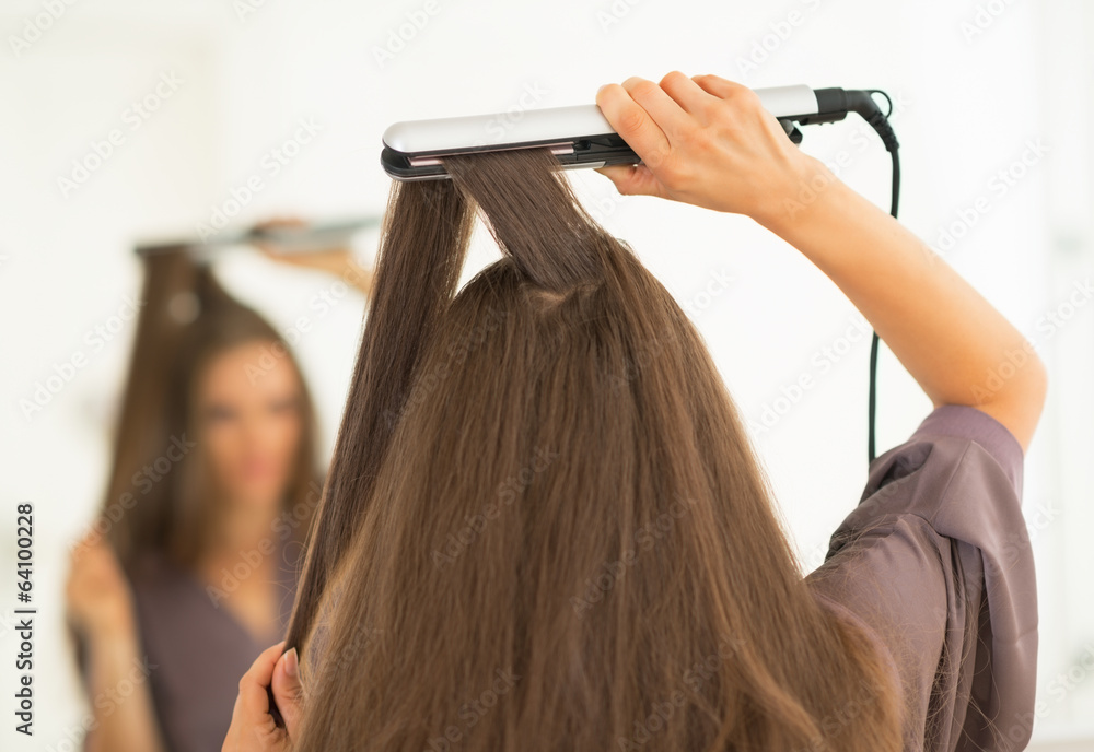 Fototapeta premium Young woman using hair straightener in bathroom. rear view